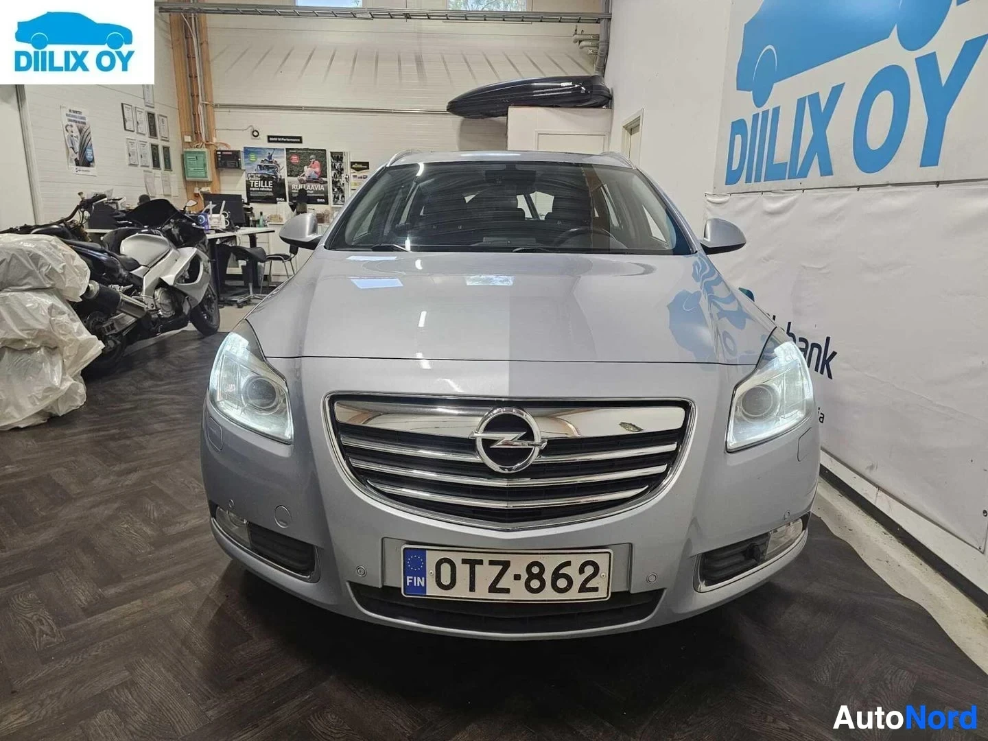 Edestä: Hopeanvärinen Opel-merkki, kuvattu etulta sisätiloissa, rekisterilevy näkyvissä