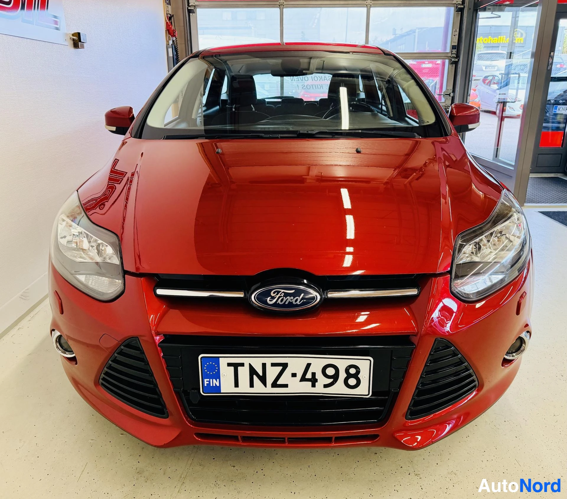 Edestä: Punainen Ford Focus, etupuoli katsottuna, Ford-tunnus keskellä, rekisterikylpi (FIN), päävalot näkyvissä, mustaväriset rengaskotelo ja säleikkö