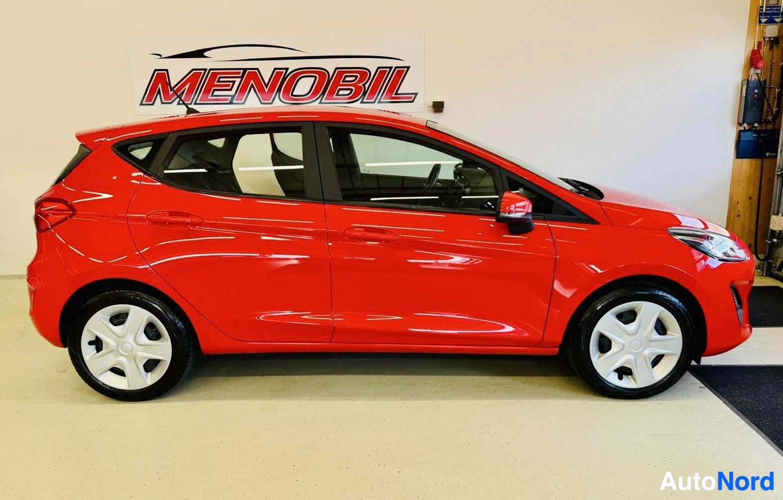 Sivulta: Punainen Ford Fiesta pysäköitynä oikealle katsottuna, näkyy koko auto sivulta. Valkoiset vanteraat, tummennetut ikkunat, showroom-tausta.