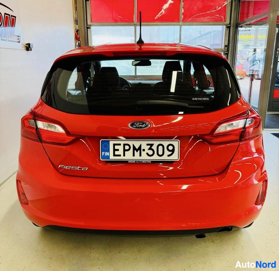 Takaa: Punainen Ford Fiesta takaa kuvattuna. Näkyy rekisterikilpi, "Fiesta" merkintä takaosassa, Ford-logo, takalampyt. Showroom-ovi taustalla