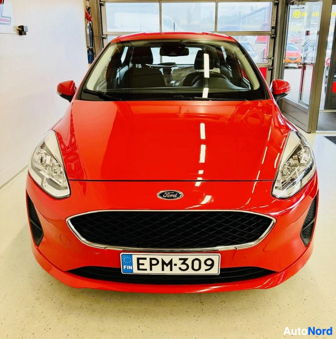 Edestä: Punainen Ford Fiesta edestä kuvattuna. Näkyy Ford-logo, tumma säleikkö, etulampyt, rekisterikilpi, punainen keula ja konepelti