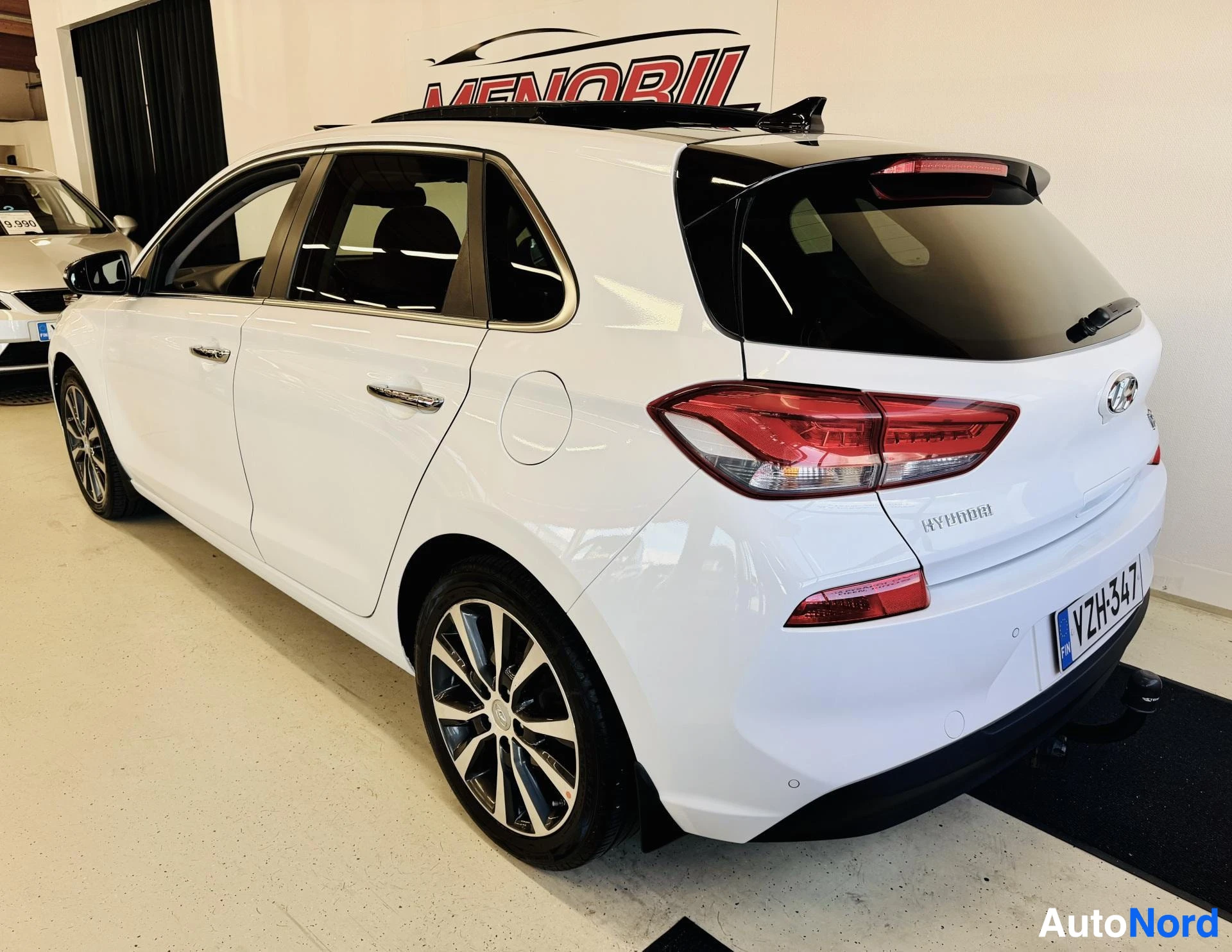 Takaviisto: Valkoinen Hyundai i30 takakolmesta oikealle kuvattuna, vasen ovi avoin, rekisterikilvellä näkyy sininen EU-merkki