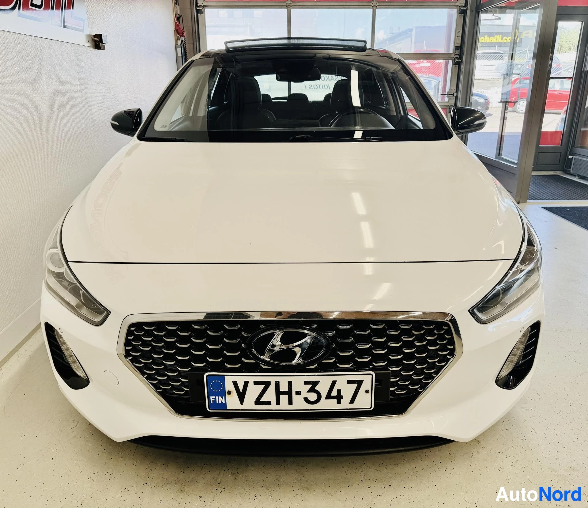 Edestä: Valkoinen Hyundai i30 edestä, rekisterinumero näkyy, mustalla silmälliisillä varustettu etusäleikköinen grilli