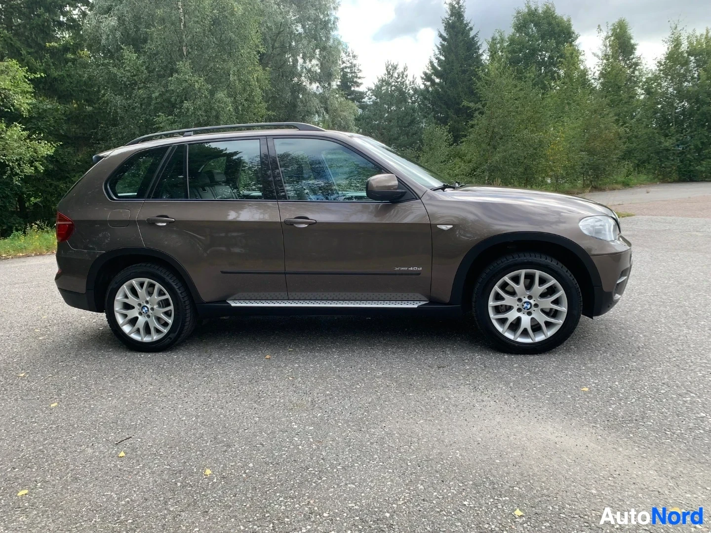 Sivulta: Ruskea BMW X5 näkyy vasemmasta sivusta, valkoised vanteet, asfalttialueella puustoa taustalla.
