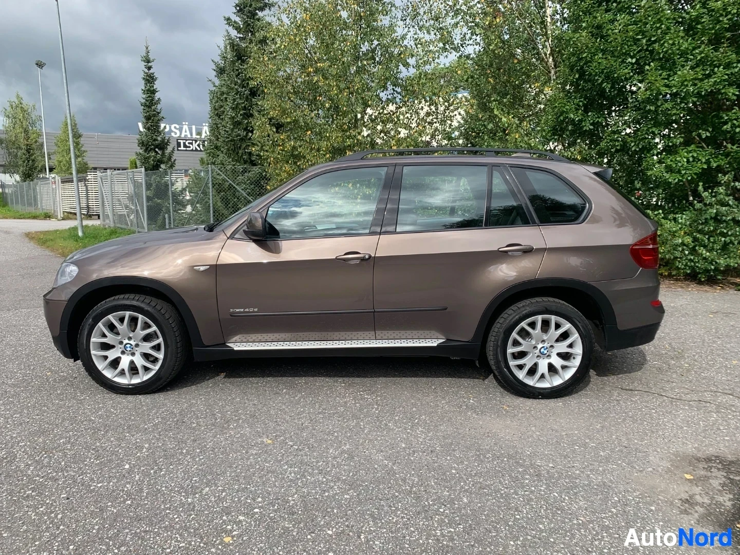 Sivulta: Ruskea BMW X5 näkyy vasemmasta sivusta, valkoised vanteet, teollisuusalue taustalla.