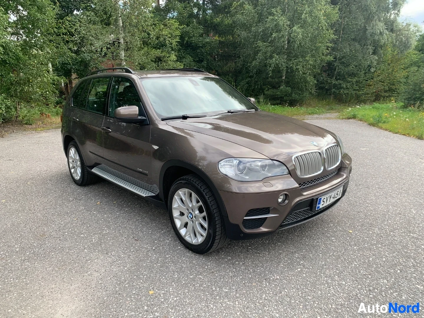 Etuviisto, nokka oikealle: BMW X5 ruskea nelivetoismaasturi sivusta oikealle, parkkipaikalla vihreän metsän edessä
