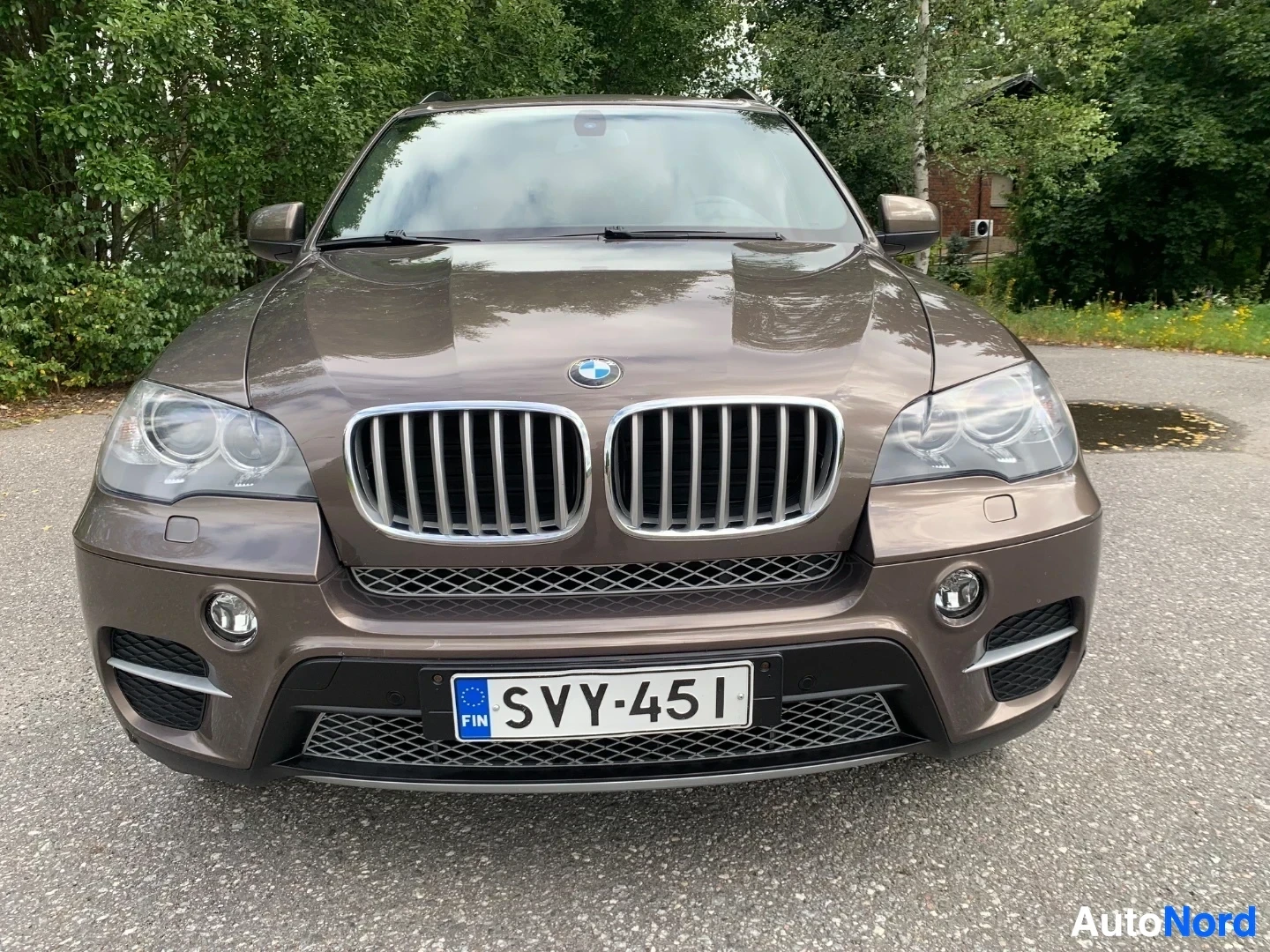 Edestä: BMW X5 ruskea SUV edestä, rekisterikilpi, vihreän puustoisen taustan edessä