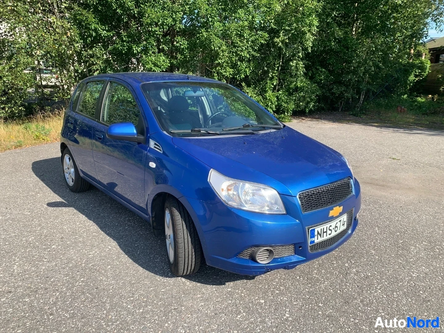 Etuviisto, nokka vasemmalle: Sininen Chevrolet Aveo -pieni tila-auto kuljettajan puolelta kolmessa neljäsosassa, sininen kattama väri, metalliset vanteet, rekisterikilvessä