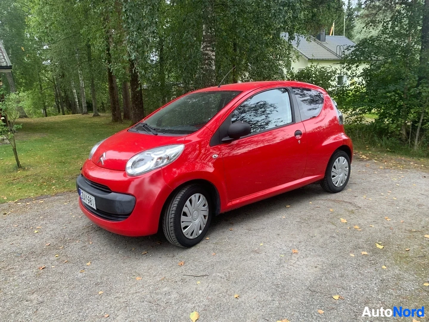 Etuviisto, nokka vasemmalle: Punainen Citroën C1 -pieni kaupunkiauto, kuvattu vasemmalta puolelta kolmen neljänneksen kulmasta, pysäköity soratiellä puiden viereen.