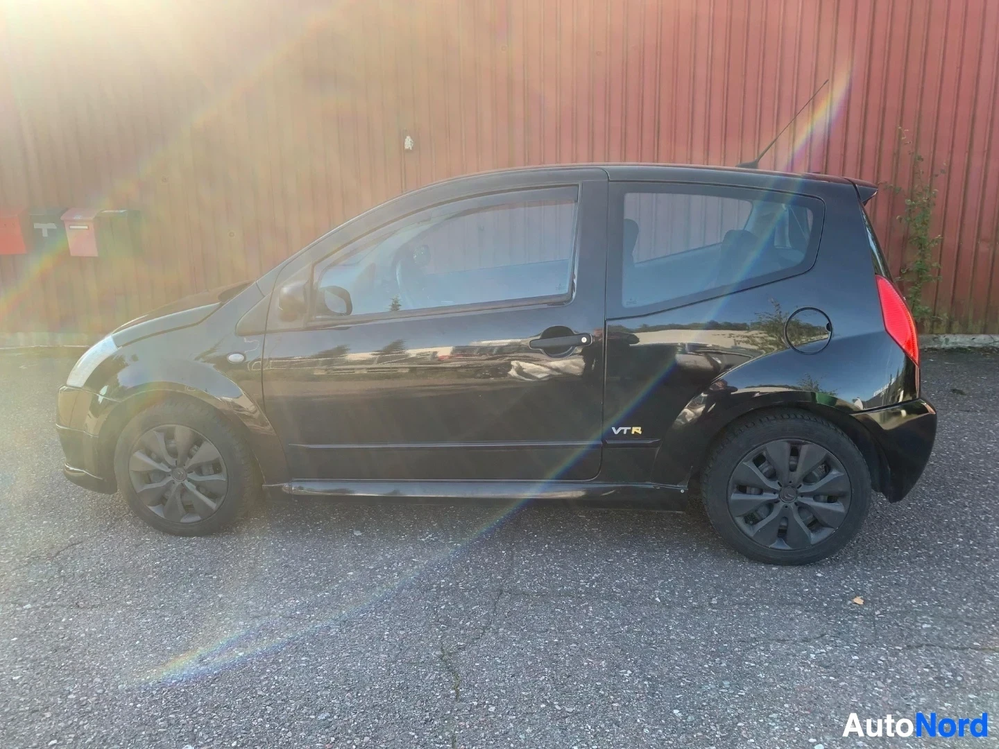 Sivulta: Musta Citroën C2 sivusta, oikealla puolella näkyvät renkaat ja osa takaosasta, punainen varasto taustalla.