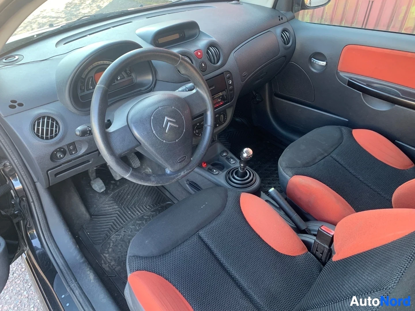 Ohjaamo: Citroën C2 sisusta, ohjauspyörä, mustat ja oranssit istuimet, musta kojelauta, mustat matot.