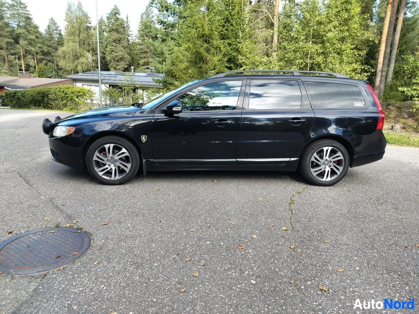 Sivulta: Musta Volvo V70 -monivaunuauto kuvattuna sivulta oikealta. Koko ajoneuvo näkyy profiilin kannalta. Suuret yksityiskohtaiset vanteet, tummuneet ikkunat, ylärulla