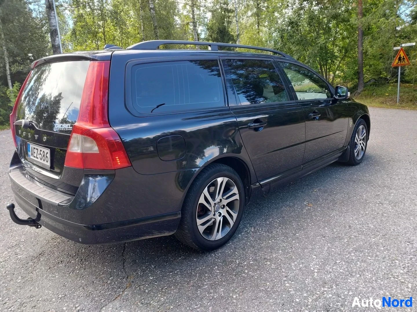Takaviisto: Musta Volvo V70 estate-auto, oikea sivu, pysäköity asfalttialueelle, puustoisella taustalla. Rekisterikyltti näkyvissä. Punainen takavalo ja hopeanväriset vante