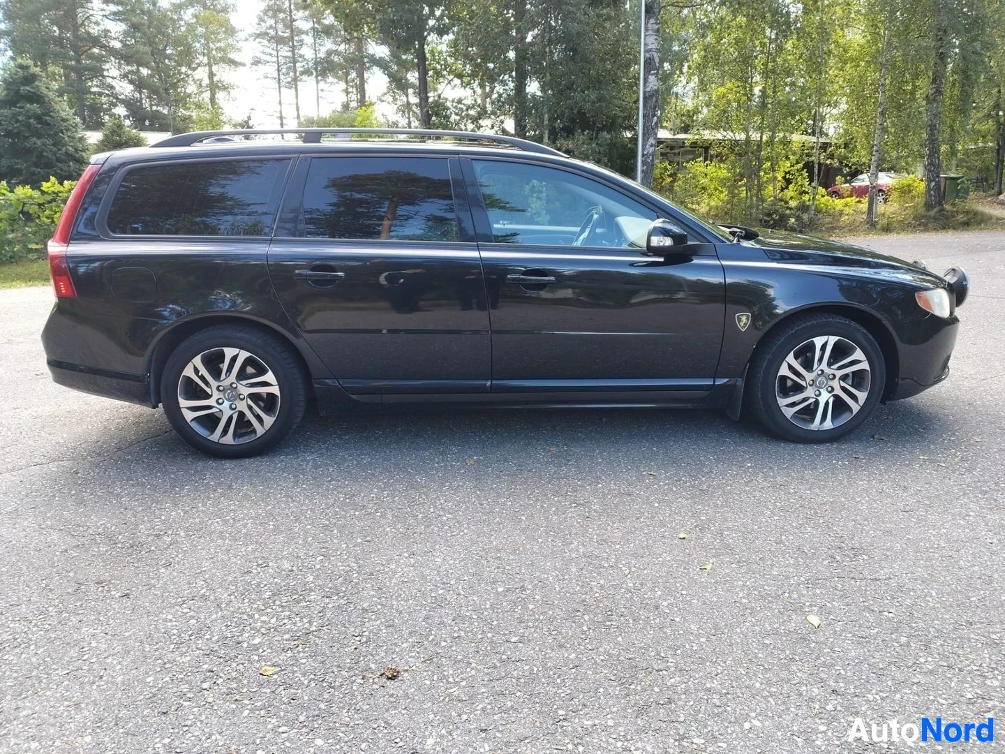 Sivulta: Musta Volvo V70 estate, vasemmanpuoleinen sivu, pysäköity asfalttialueelle. Hopeanväriset vanteet, tummennnetut ikkunat, Volvo-merkki näkyvissä. Taustalla puita