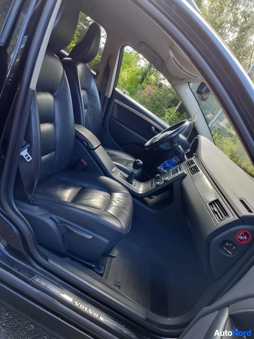 Ohjaamo: Volvo V70:n etukabiinikuva, oikea puoli, oven avautuessa näkyvät mustat nahkaistuimet, monikäyttöinen ohjain, konsolilla sininen näyttö ja punainen painike, ovi