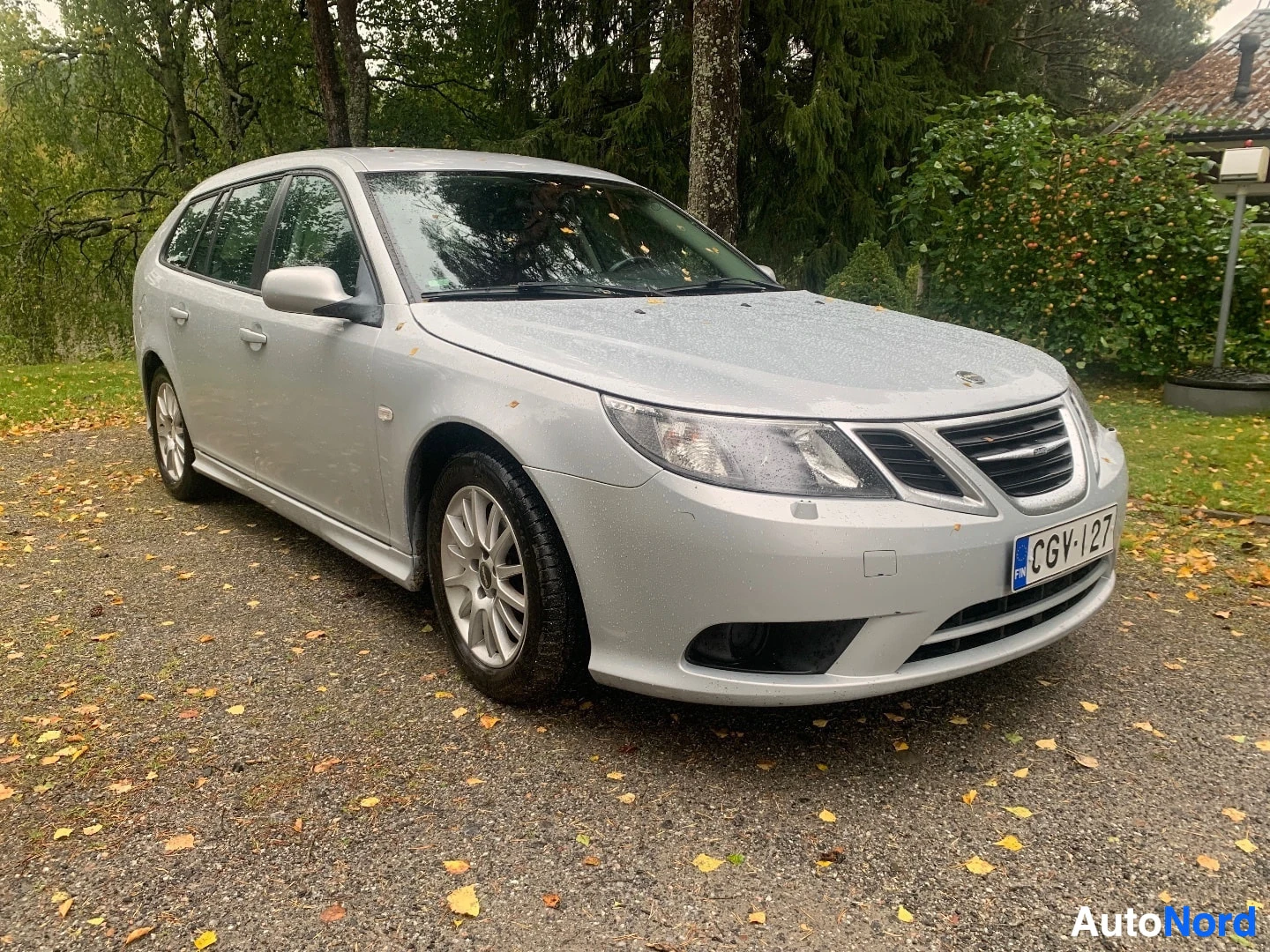 Etuviisto, nokka oikealle: Hopeanvärinen farmariauto, Saab 9-3, kuvattuna edestä oikealta. Autossa on valkoiset rekisterikilvet tekstillä ja edessä pyöreät sumuvalot. Pysäköity s