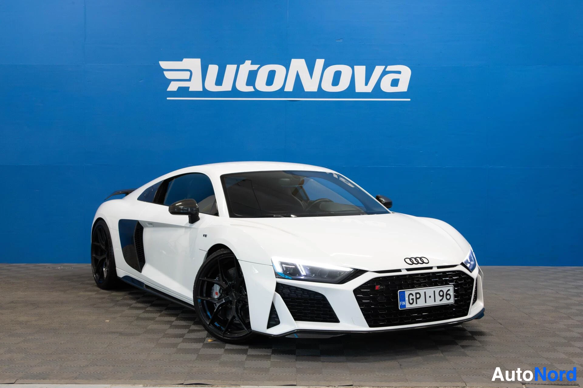 Etuviisto, nokka oikealle: Valkoinen Audi R8, jossa on mustat vanteet, pysäköitynä viistosti oikealle etuviistoon sinisen seinän edessä. Rekisterikilpi näkyy. Valot ovat päällä.