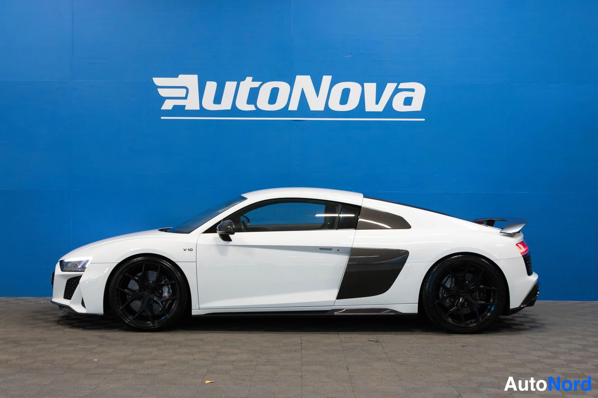 Sivulta: Valkoinen Audi R8 V10 sivusta, koko ajoneuvo näkyvissä, mustat alloy-vanteet, AutoNova-tausta, sininen seinä
