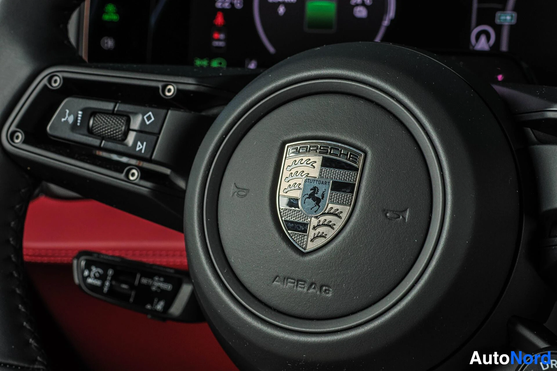 Ohjaamo: Lähikuva ohjauspyörästä, jossa on Porsche-logo keskellä ja monitoiminäppäimet vasemmalla puolella. Mittaristo näkyy epätarkkana taustalla.