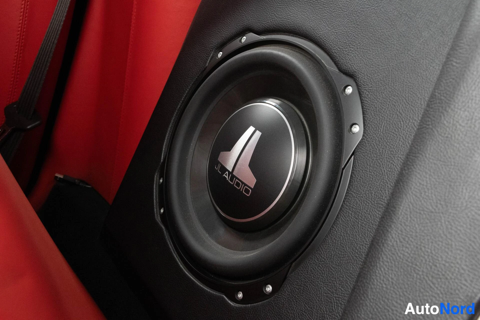 Yksityiskohta: JL Audio -subwoofer autossa. Kaiuttimessa lukee "JL AUDIO" ja "JL". Punaiset istuimet näkyvät taustalla.