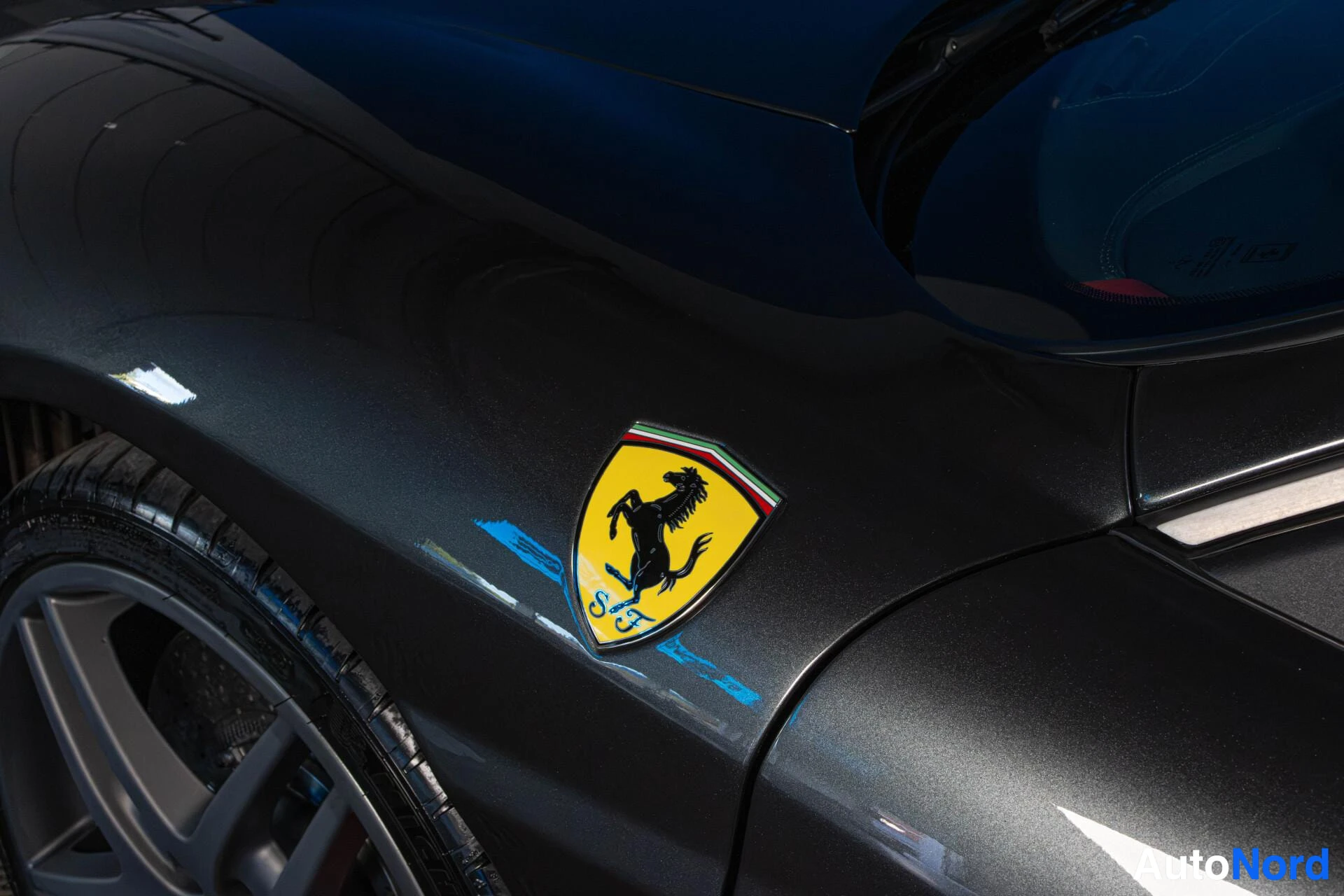 Yksityiskohta: Lähikuva harmaan urheiluauton etuosasta, jossa näkyy Ferrari-logo ja osa etupyörästä.