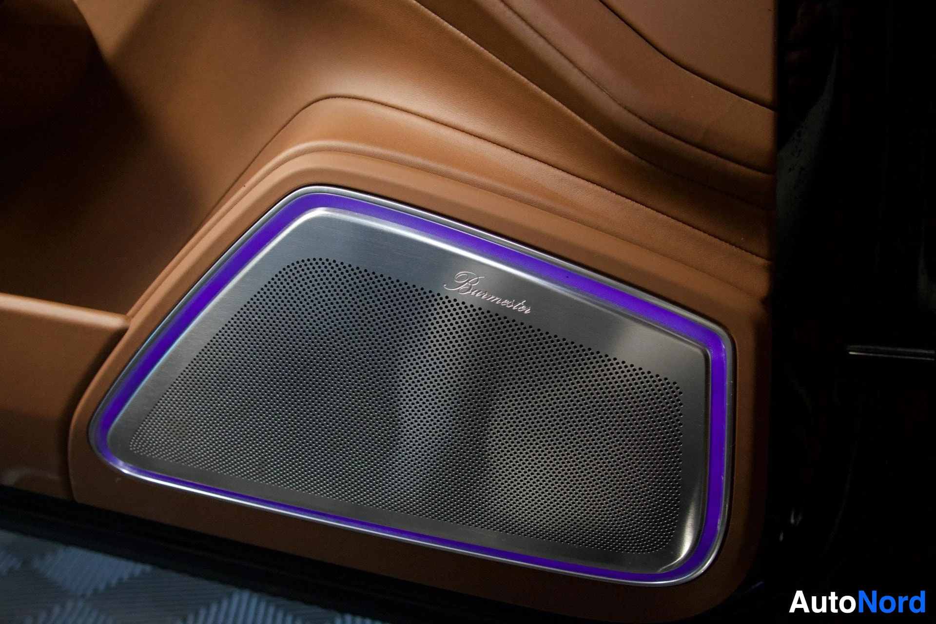 Yksityiskohta: Lähikuva auton sisätilojen kaiuttimen ritilästä, jossa on "Burmester"-logo ja violetti tunnelmavalaistus.