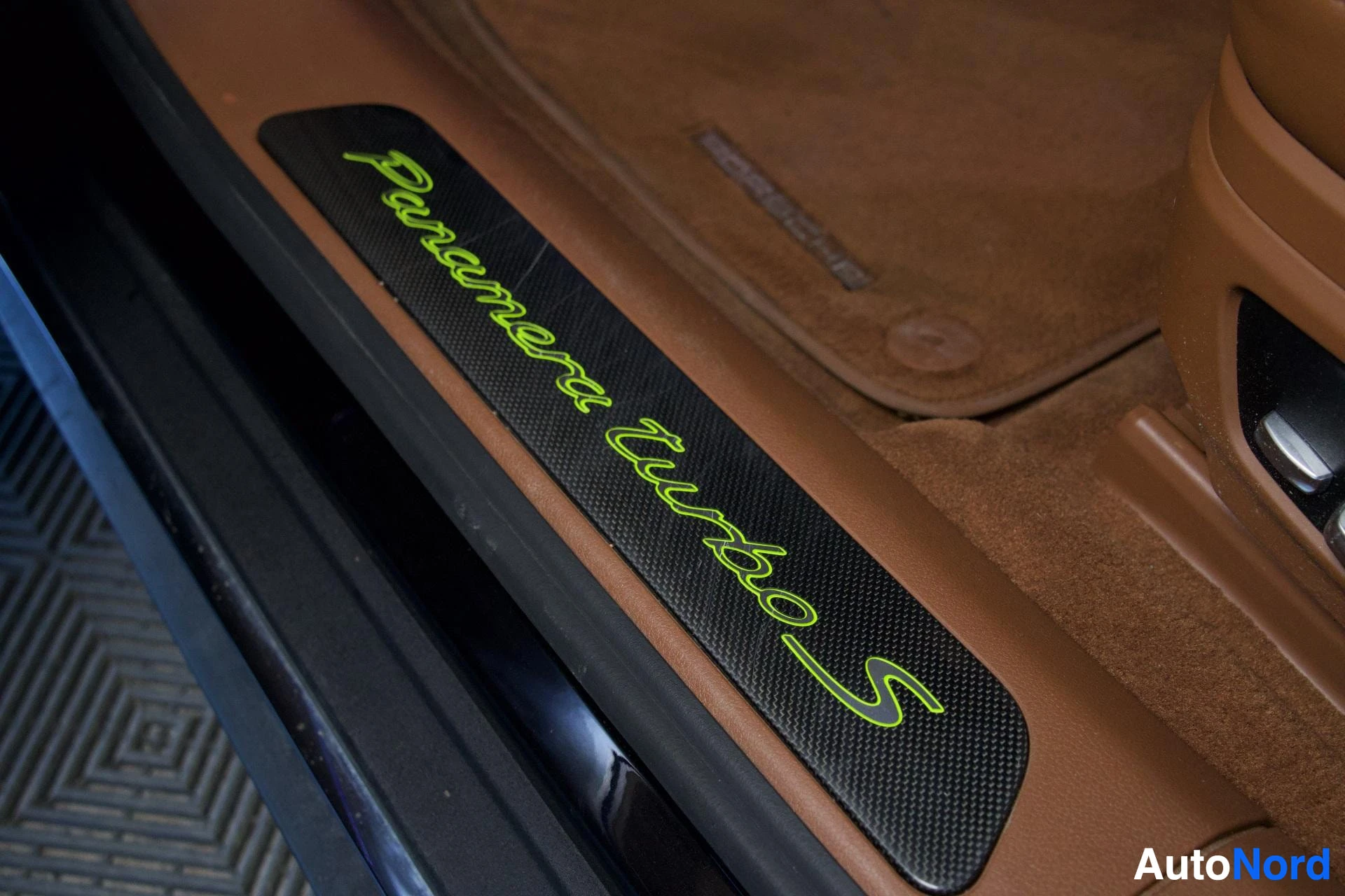 Yksityiskohta: Lähikuva oven kynnyslistasta, jossa lukee "Panamera Turbo S" vihreällä tekstillä mustalla hiilikuitupohjalla, ruskeaa mattoa ja auton sisustusta vasten.