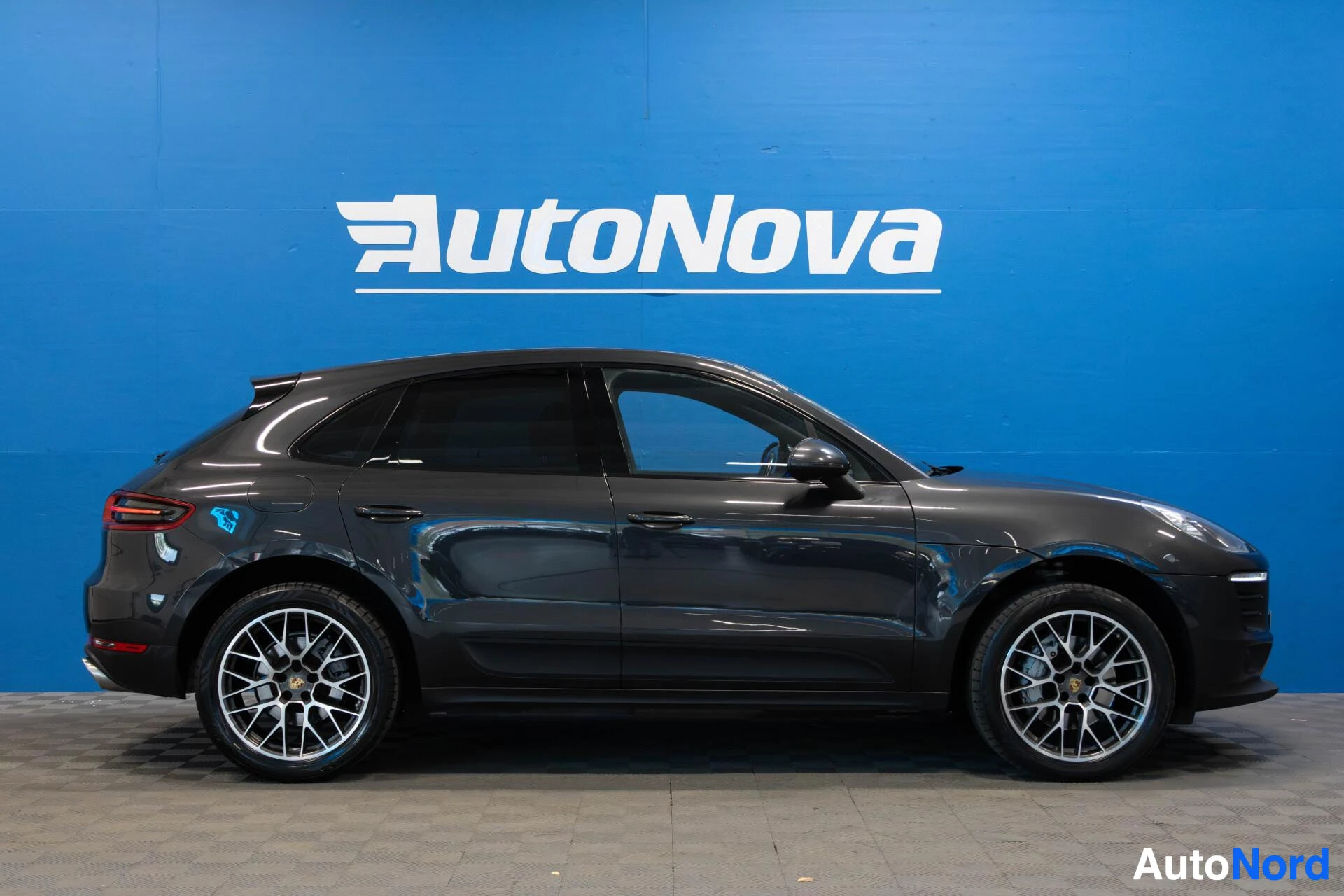 Sivulta: Sivukuva tummanharmaasta Porsche Macan -autosta, jossa on auton sivu sinistä AutoNova-seinää vasten, mustat ikkunat ja monipuolaiset kevytmetallivanteet.
