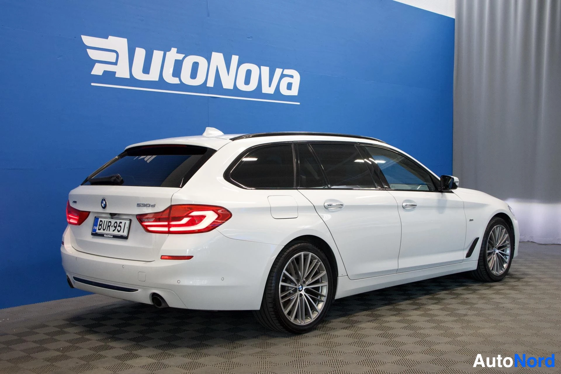 Takaviisto: Valkoinen farmariauto kuvattu takaviistosta oikealta puolelta. Auton takavalot ovat päällä. Takakontissa on mallimerkintä "530d" ja BMW-logo. Rekisterikilvessä