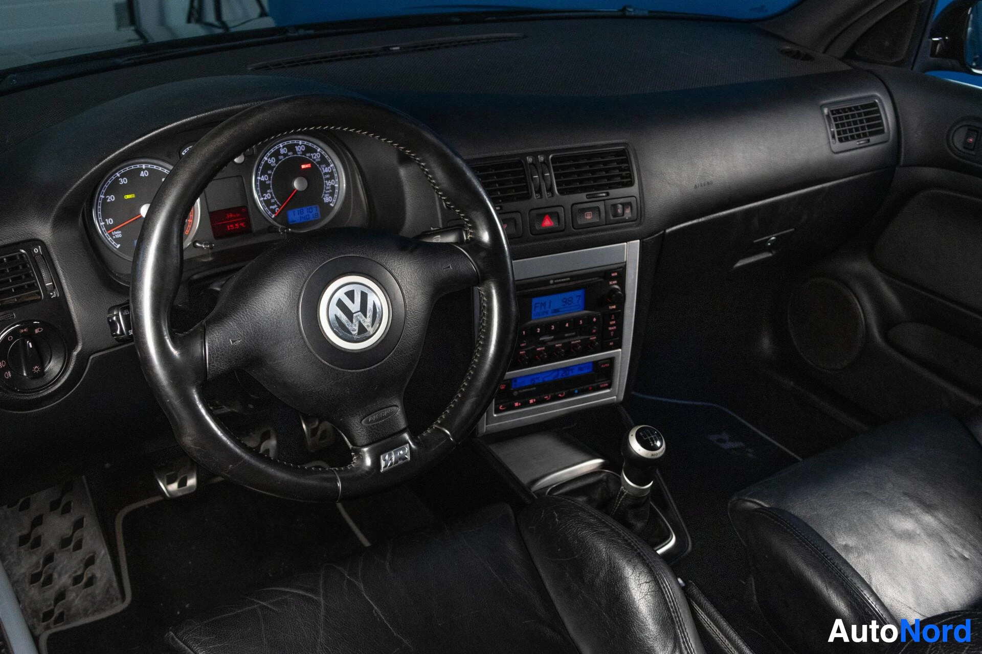 Etuistuimet: Auton kuljettajan istuimen näkymä: Volkswagen-ohjauspyörä, kojelauta mittareineen ja sininen näyttö, mustat nahkaiset istuimet, käyttökytkimet ja ilmanvaihtokau