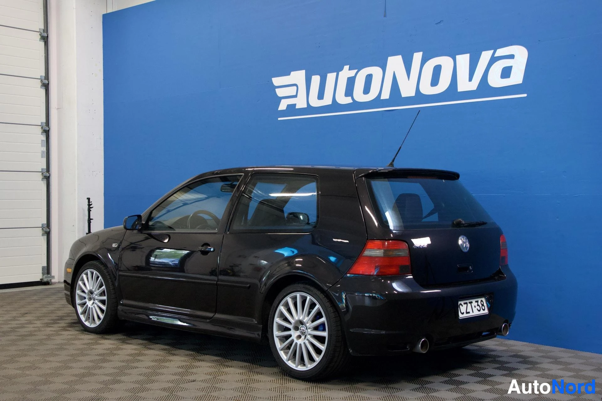 Takaviisto: Musta Volkswagen Golf pysäköitynä profiilinäkymässä oikealle päin, AutoNova-huoltohallin sinistä seinää vasten. Kuljettajan ovi on avoinna. Valkoiset vanteet, p