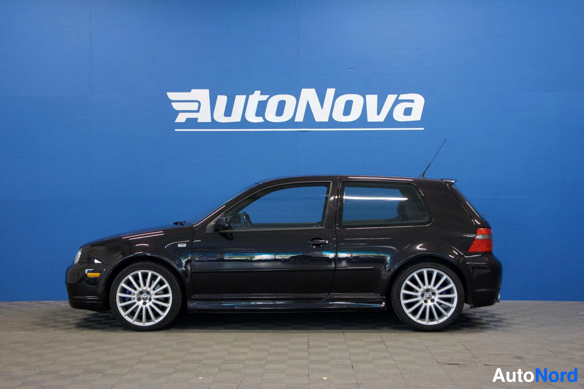 Sivulta: Musta Volkswagen Golf kuvattu sivusta vasemmalle suuntautuvassa profiileissa AutoNova-hallissa sinisen seinän edessä. Valkoiset vanteet, tummennetut ikkunat, si