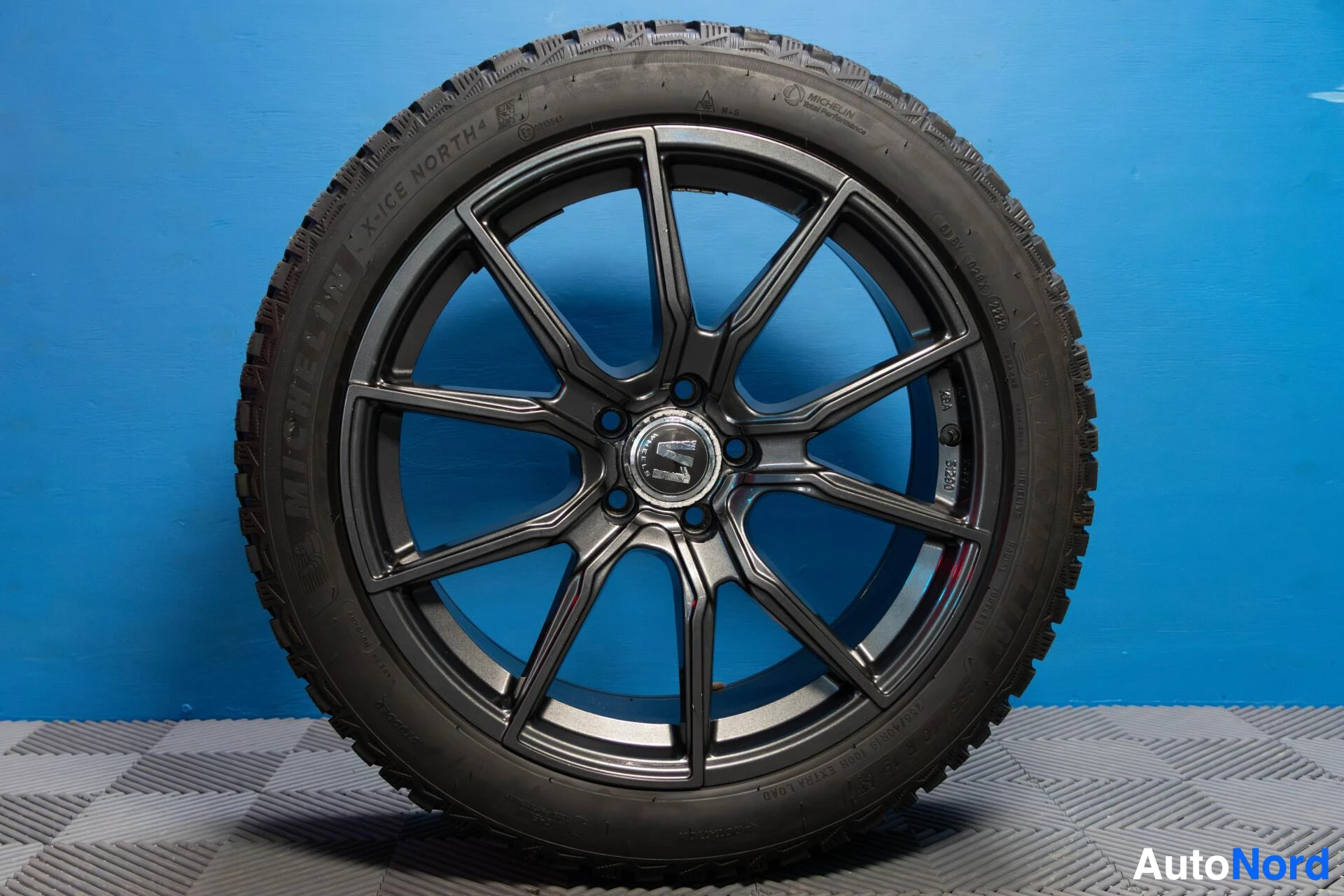 Yksityiskohta: Lähikuva mustasta kevytmetallivanteesta, jossa on Michelin X-ICE NORTH 4 -merkitty nastarengas. Vanteessa on keskellä valmistajan logo.