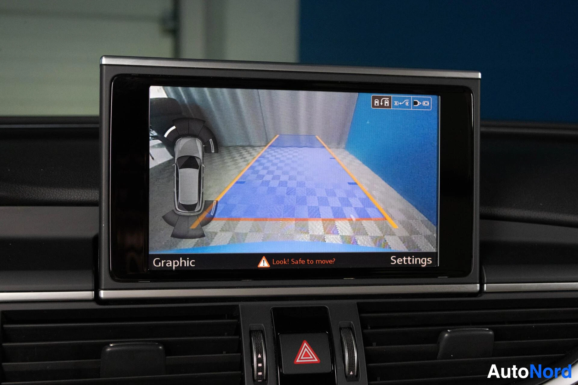 Ohjaamo: Kuva auton infotainment-näytöstä, jossa näkyy peruutuskameranäkymä ja auton yläpuolinen 360 asteen näkymä ohjauslinjoilla. Näytön alareunassa lukee "Graphic", "