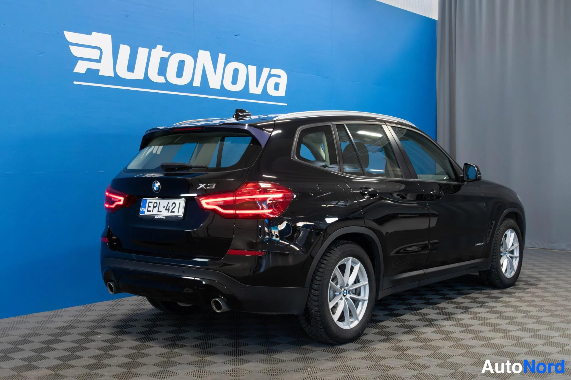 Takaviisto: Musta BMW X3 näkyy takaa kolmen neljänneksen kulmasta AutoNova-kaupan siniseen seinään nojaten. Näkyviin kirjaimet rekisteriluvusta alkavat