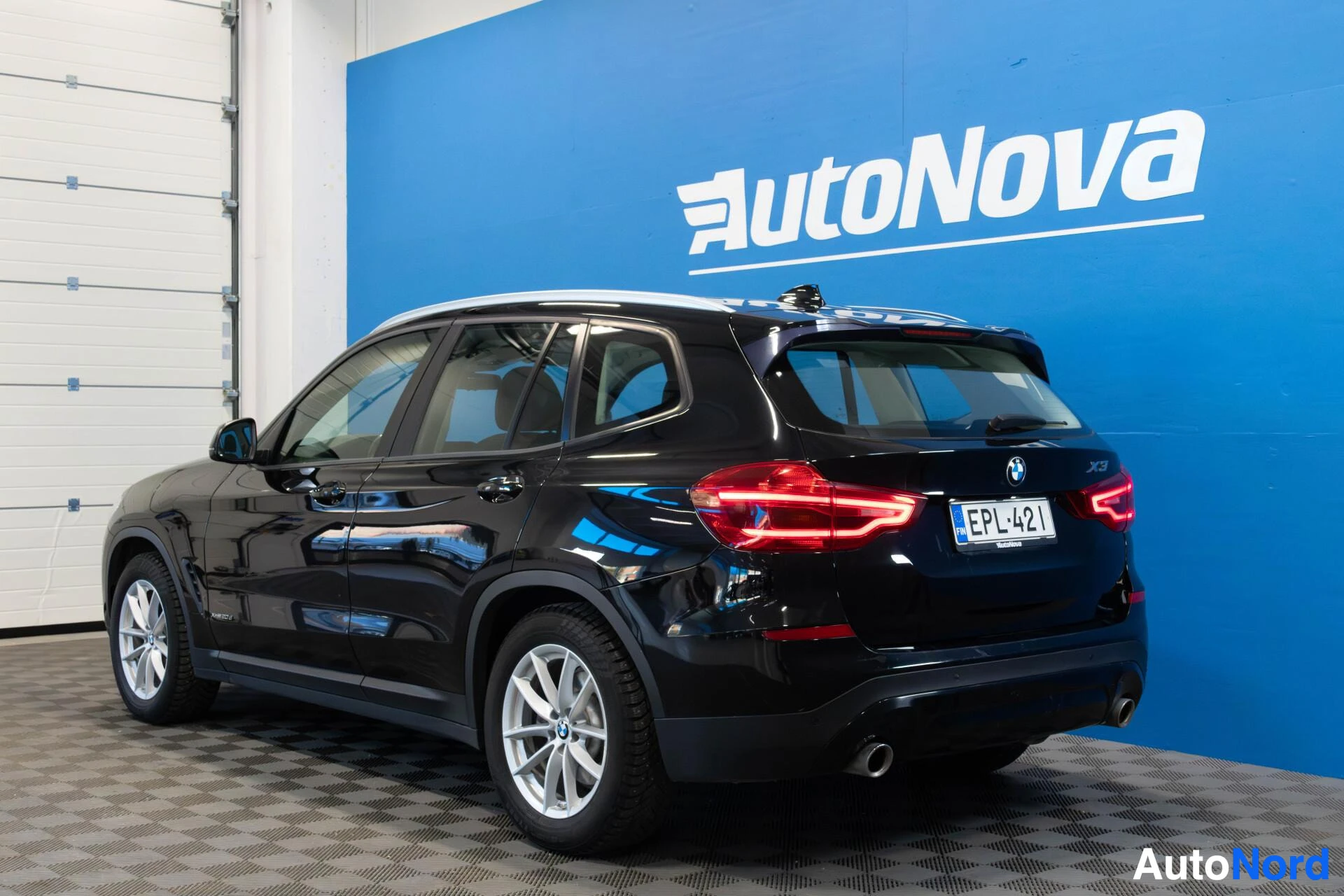 Takaviisto: Musta BMW X3 näkyy takaa kolmella neljäsosalla, valkoisen autohallin ja sinisen AutoNova-kyltin edessä. Rekisterikilpi näkyy. Valkokalliset vantereet ja