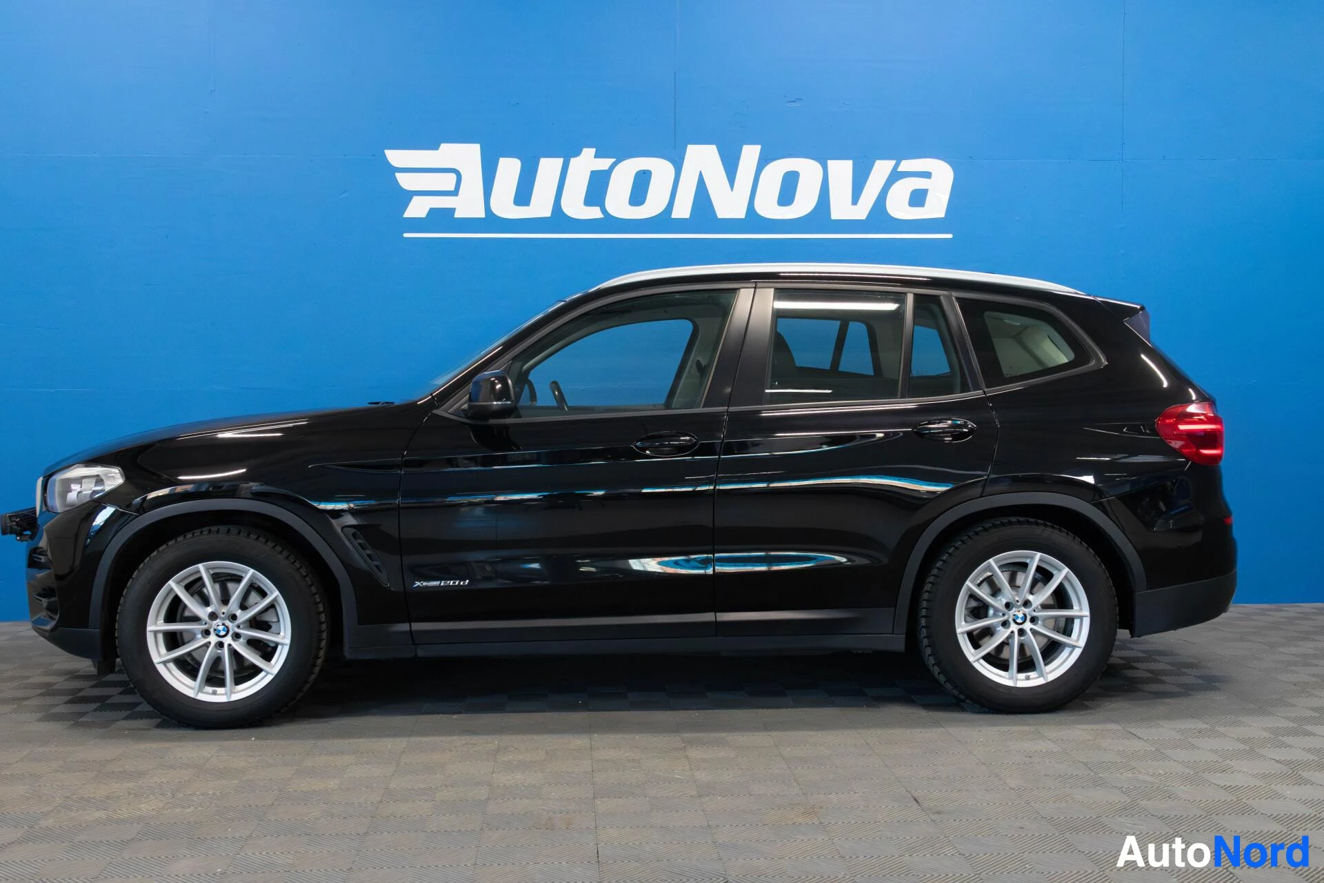 Sivulta: Musta BMW X3 näkyy sivulta vasemmalta, sinisen AutoNova-kyltin edessä. Valkokalliset vantereet, mustat ikkunat ja senkaltaiset osat näkyvät selvästi.