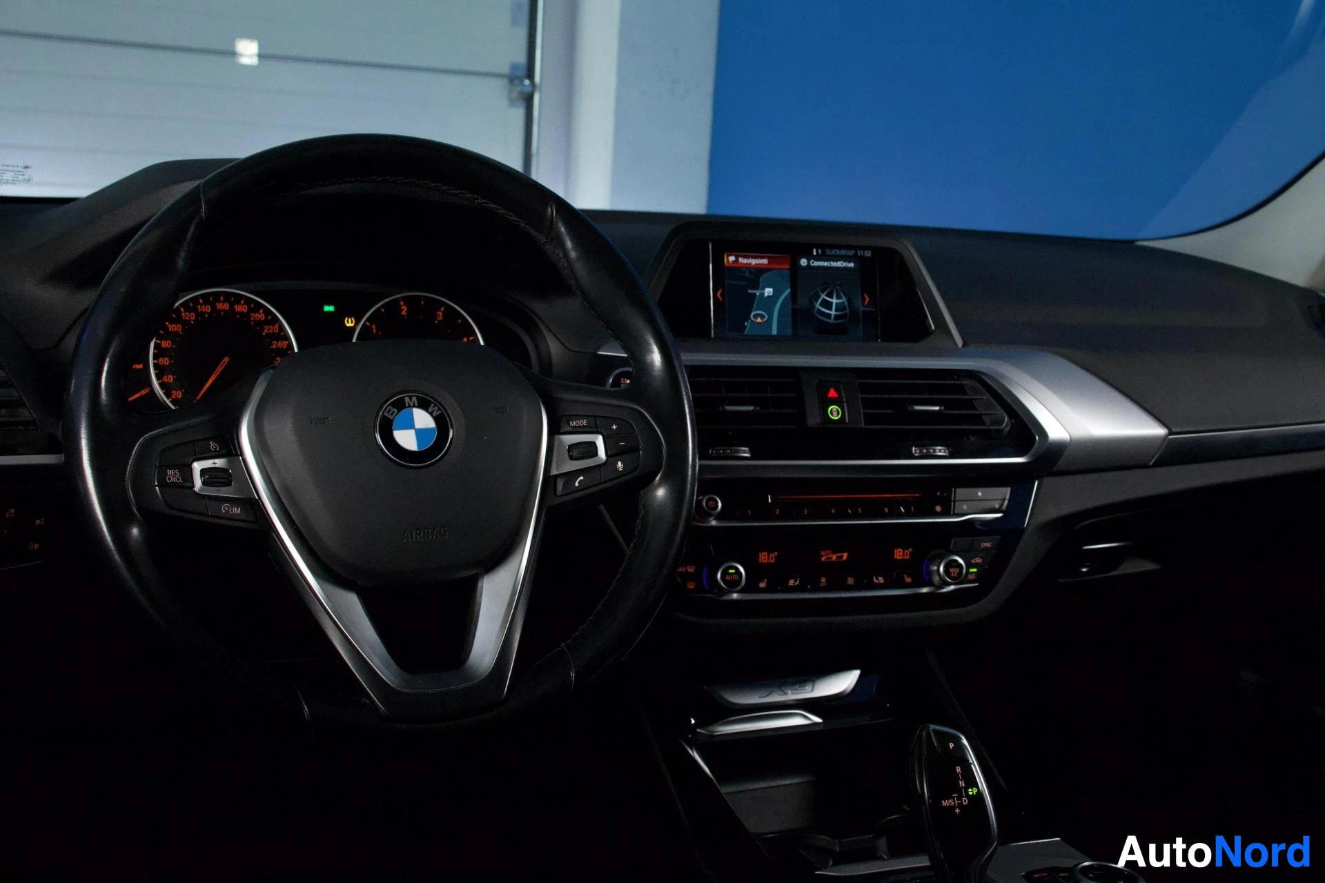 Etuistuimet: Auton sisustus etulta: musta ratti BMW-logolla, digitaalinen värinäyttö, ilmastoinnin säätimet ja keskikonsoli sinisellä taustalla.