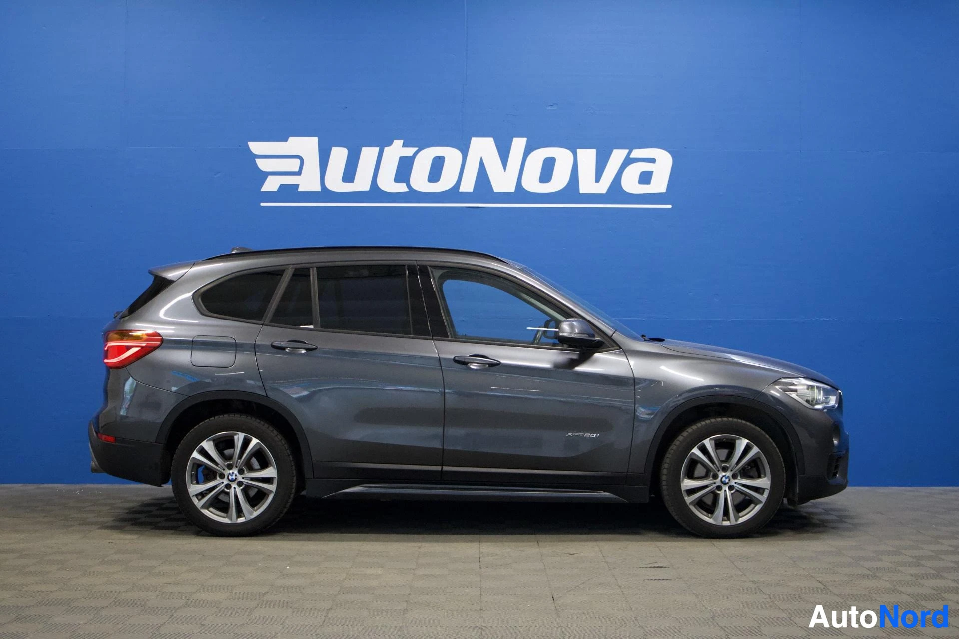 Sivulta: Sivuprofiili harmaasta BMW X1 -autosta, joka on pysäköity autonäyttelytilan sinisen seinän eteen. Autossa on kattokaiteet ja kevytmetallivanteet.