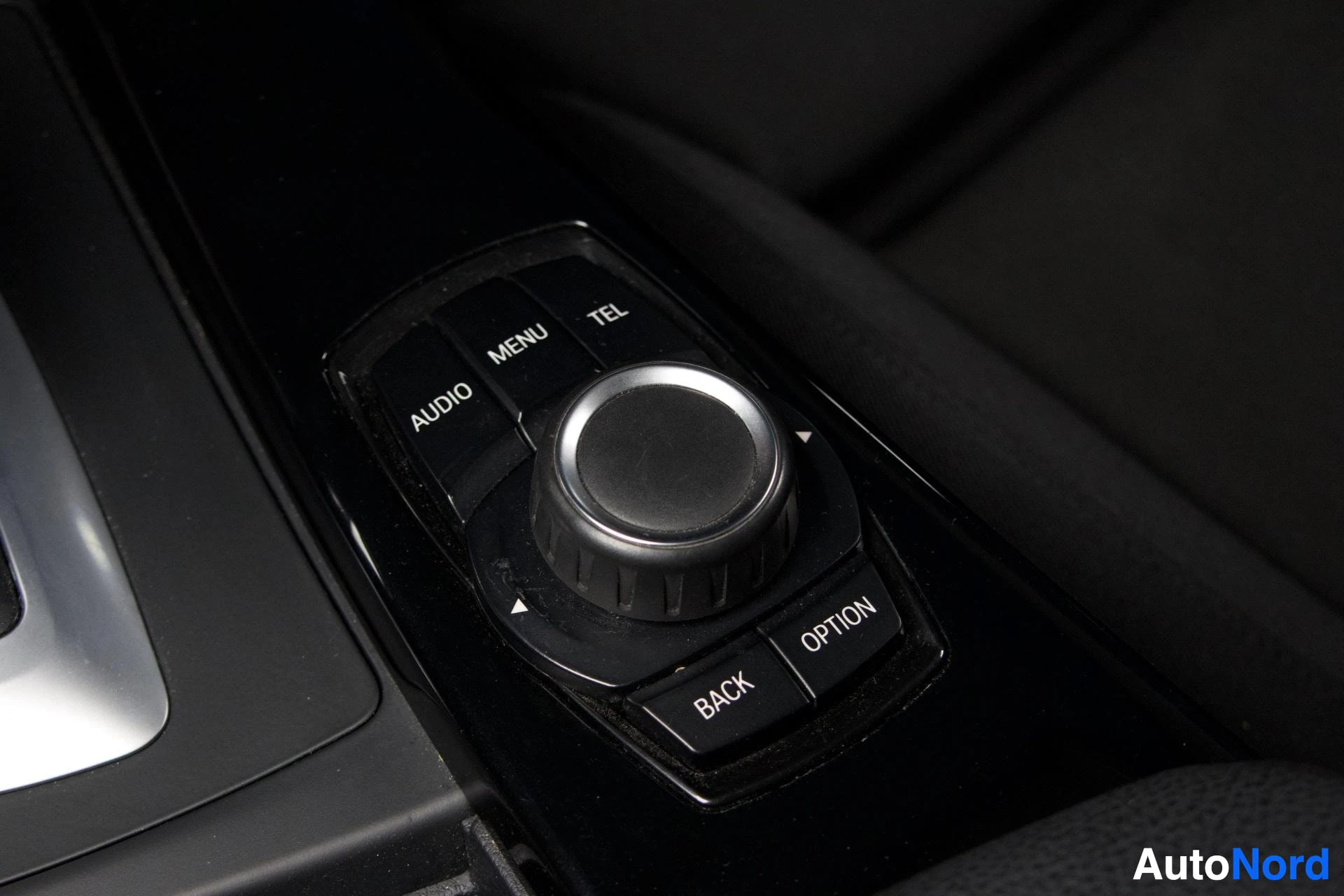 Yksityiskohta: Lähikuva ohjaamon mustasta iDrive-joystick-ohjainpyörästä tekstillä AUDIO, MENU, TEL, OPTION ja BACK painikkeilla, hopeanvärinen pääkehä, musta sisusta taustall