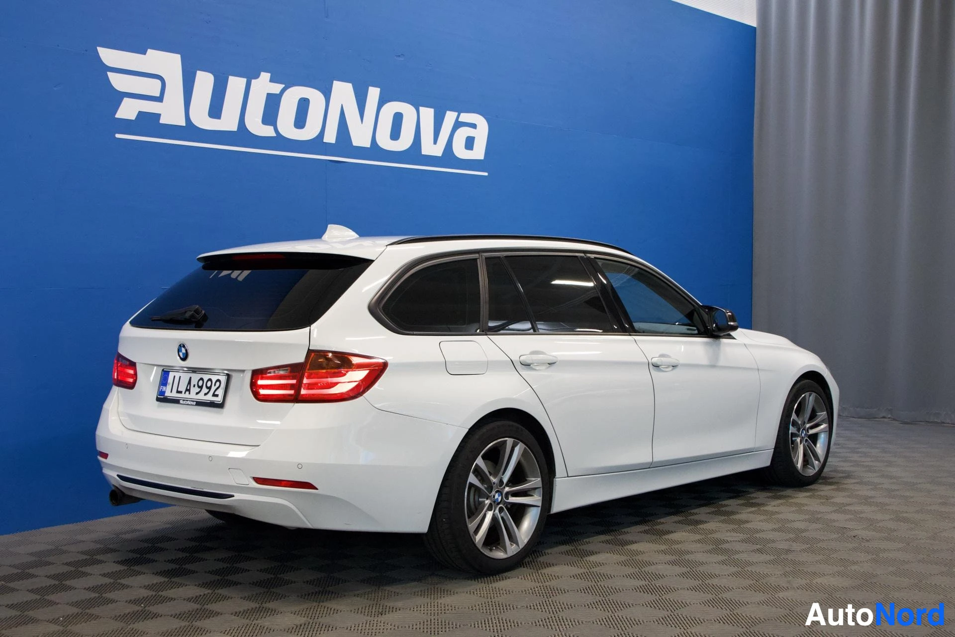 Sivulta: Valkoinen BMW-farmari näkyy sivusta oikealla, sinisen AutoNova-seinän edessä