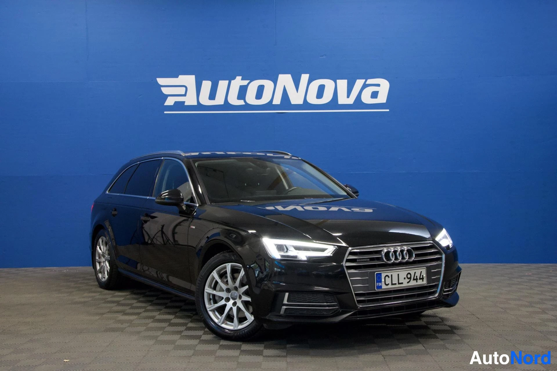 Etuviisto, nokka oikealle: Musta Audi A4 Avant, jossa on kattokaiteet ja alumiinivanteet, seisoo suoraan edestäpäin kuvattuna AutoNova-logon edessä. Ajovalot ovat päällä.