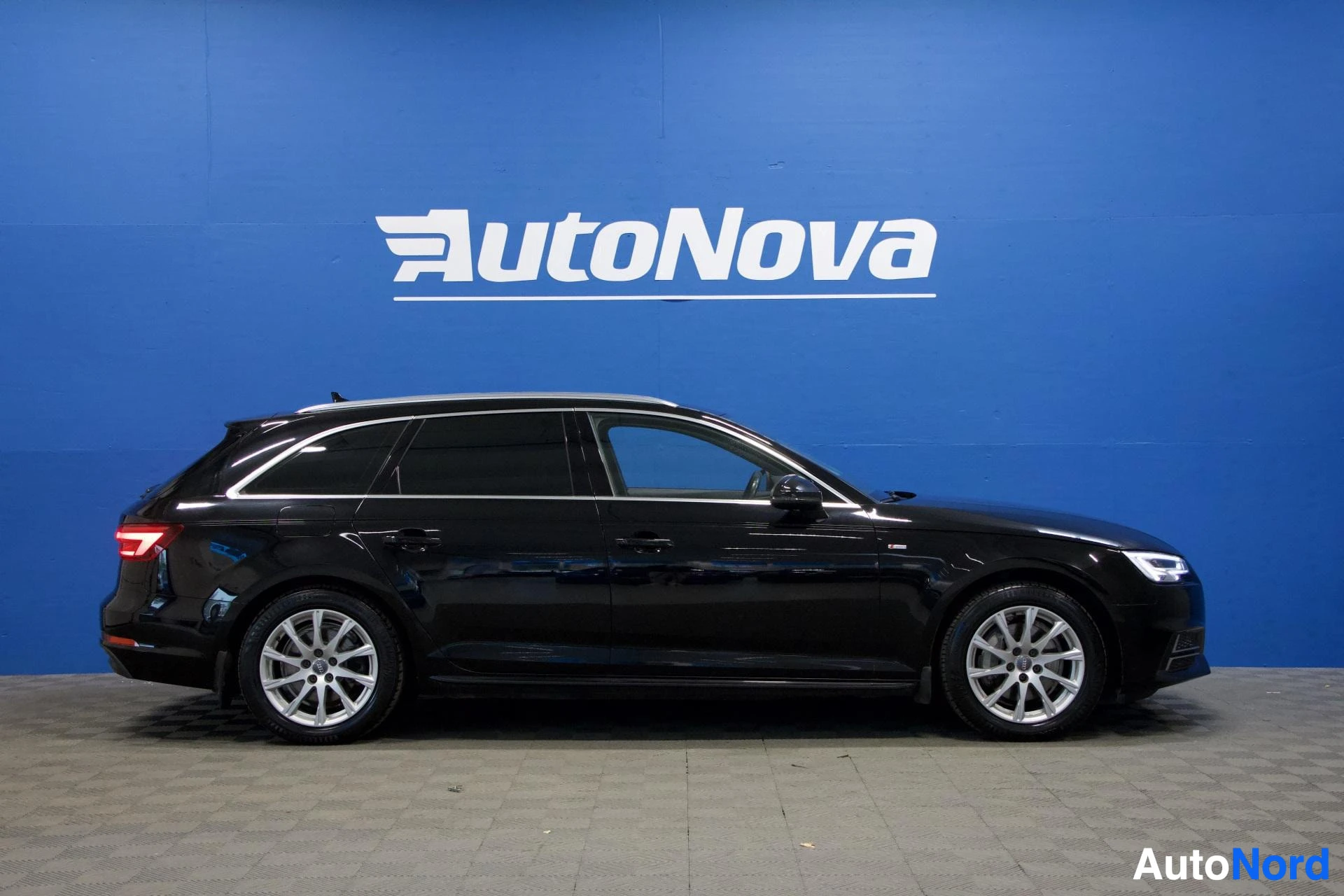 Sivulta: Musta Audi A4 Avant seisoo sivusta katsottuna sinisen AutoNova-seinän edessä.