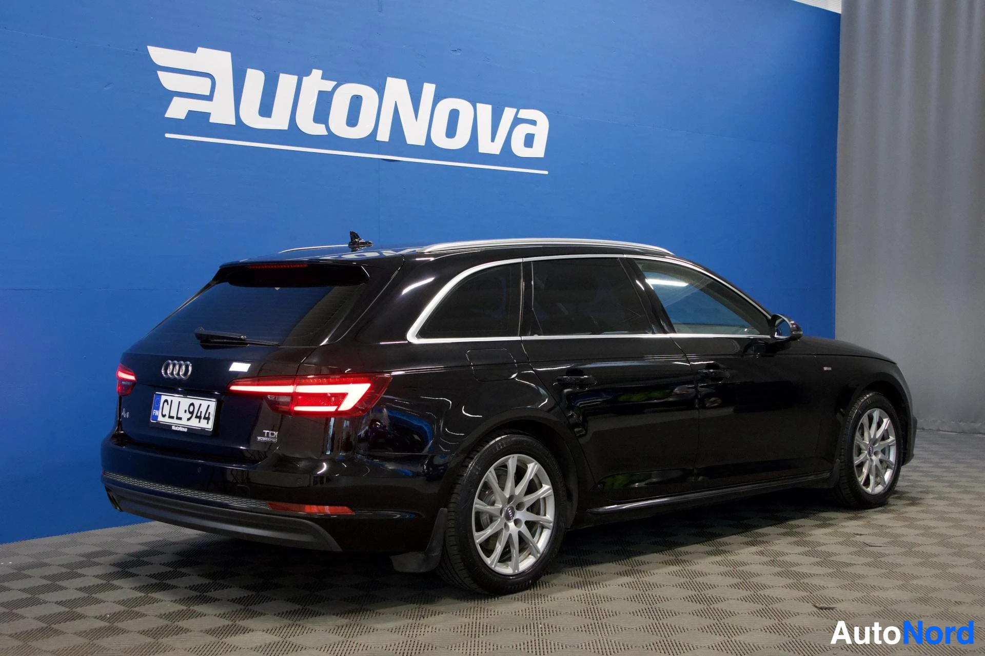 Takaviisto: Musta Audi-farmari nähdään oikealta sivulta takaapäin. Ajoneuvo on pysäköity sinisen AutoNova-seinän eteen. Rekisteritunnus on näkyvissä takalaidassa. A