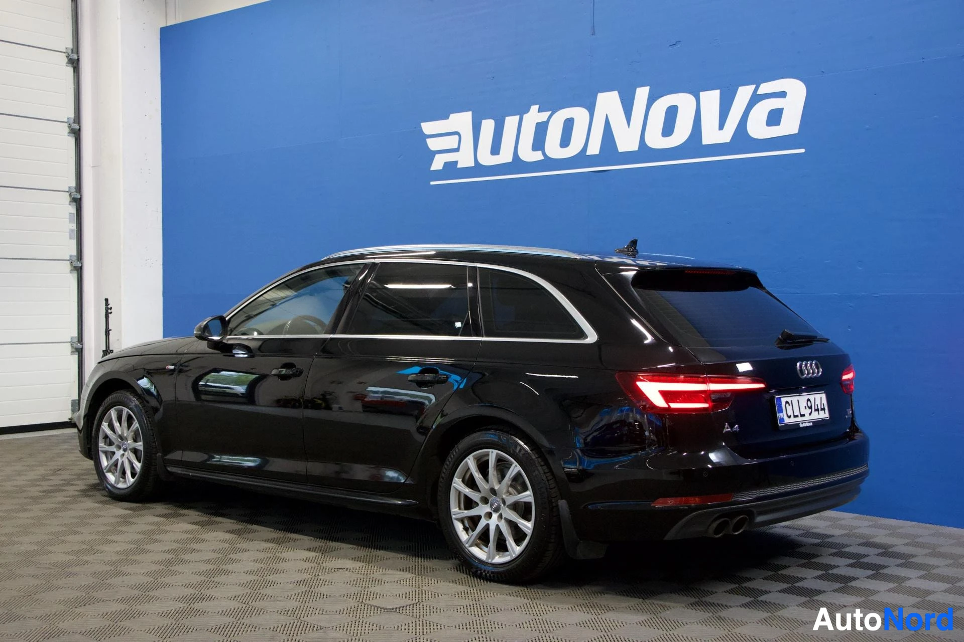 Takaviisto: Musta Audi A4 Avant seisoo sinisen AutoNova-seinän edessä, kuvattu oikealta puolelta taakse päin. Näkyy rekisterikyltti, valkolakat, LED-takavalot ja takaovi.