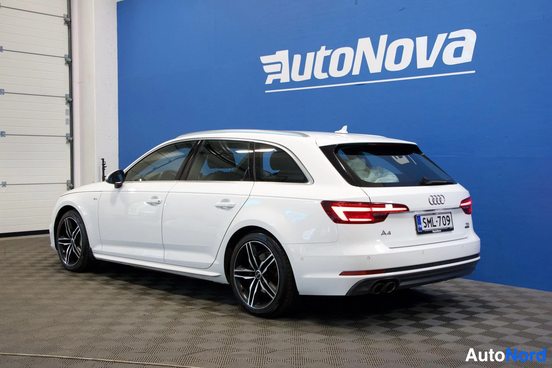Takaviisto: Valkoinen Audi A4 Avant -farmari kylhelta kuvattuna autoliikkeen sinisen seinän edessä, valkoinen seinä ja betonilattiaa vasemmalla, rekisterikilpi näky