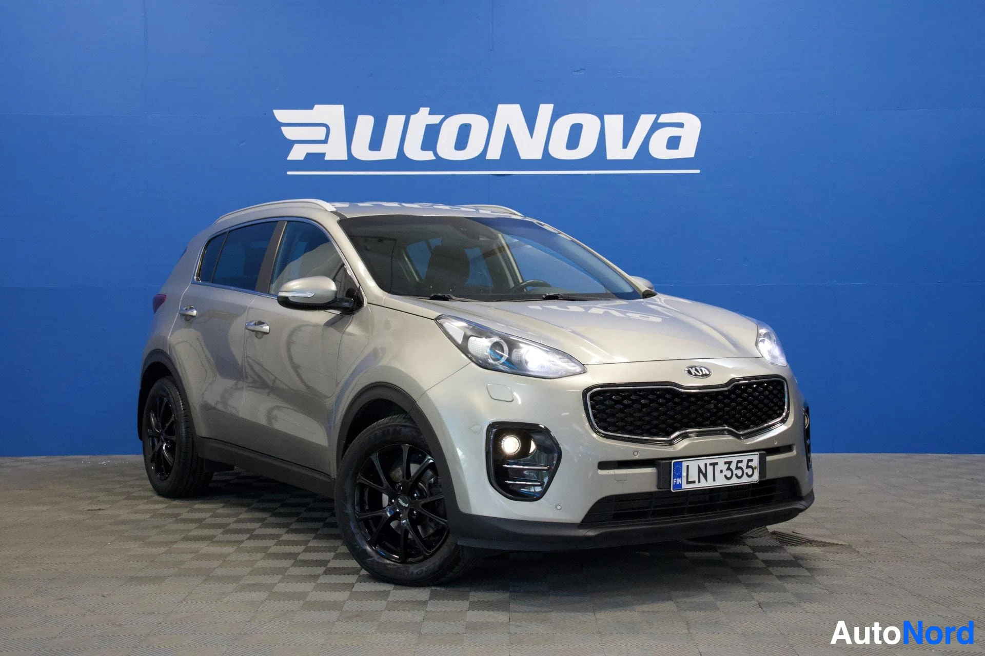 Etuviisto, nokka oikealle: Beigenvärinen Kia Sportage SUV edestä ja vasemmalta sivulta kuvattuna. Autossa on mustat monipuolaiset kevytmetallivanteet ja katolla kattokaiteet. Rekis