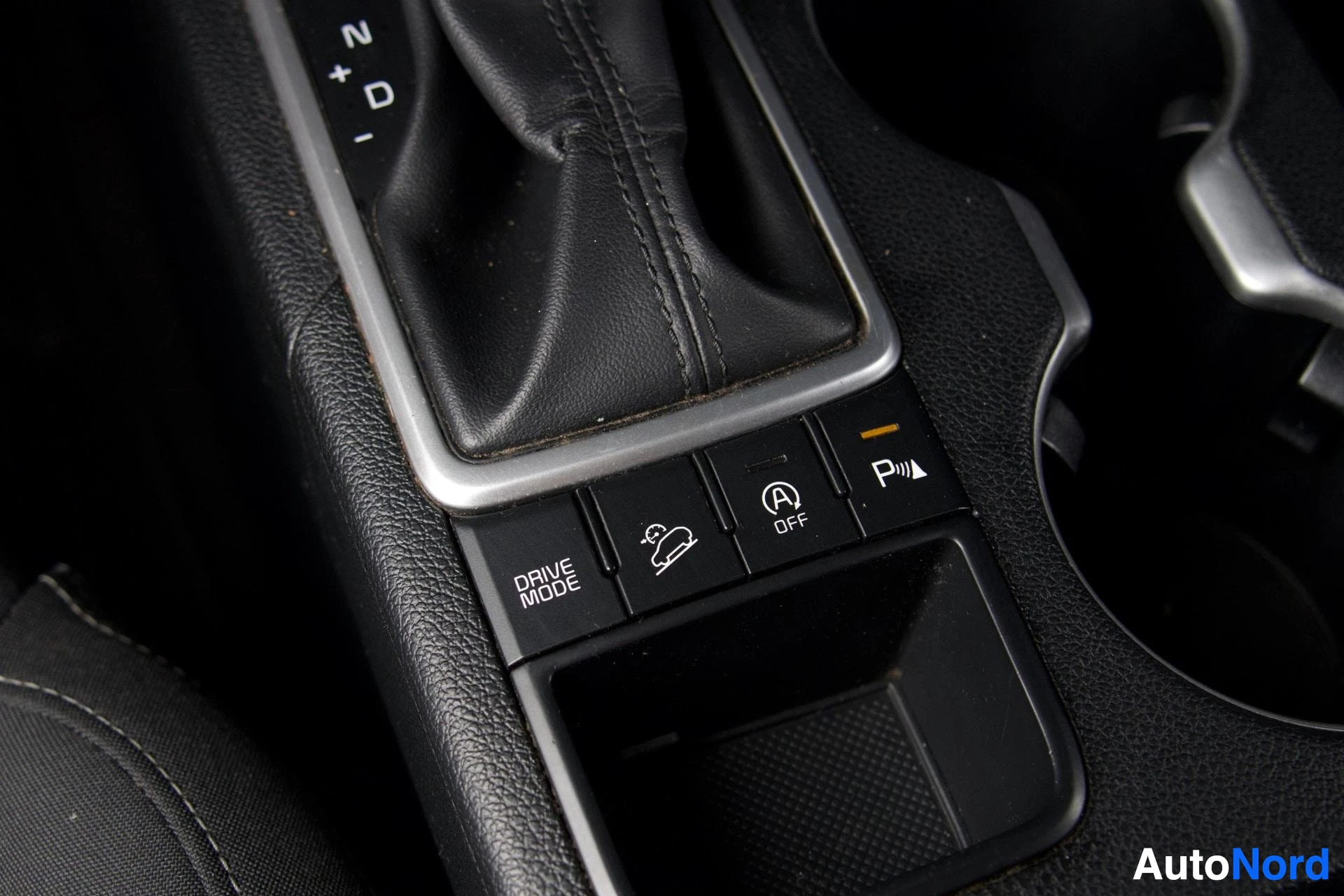 Yksityiskohta: Lähikuva auton keskikonsolista, jossa näkyy vaihdekeppi ja painikkeita. Painikkeissa lukee "DRIVE MODE", "OFF" ja pysäköintitutkan symboli. Yksi painikkeista on