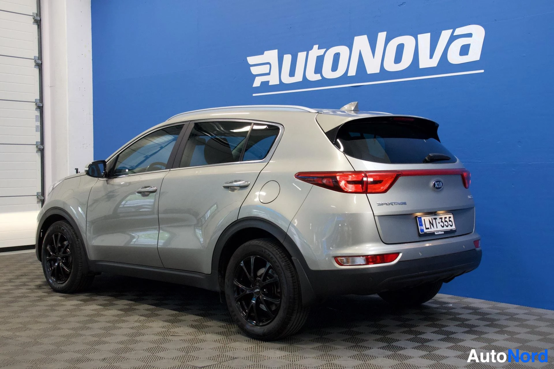 Takaviisto: Harmaa Kia Sportage kuvattuna takavasemmalta AutoNova-logon edessä. Ajoneuvossa on mustat kevytmetallivanteet ja takarekisterikilpi.
