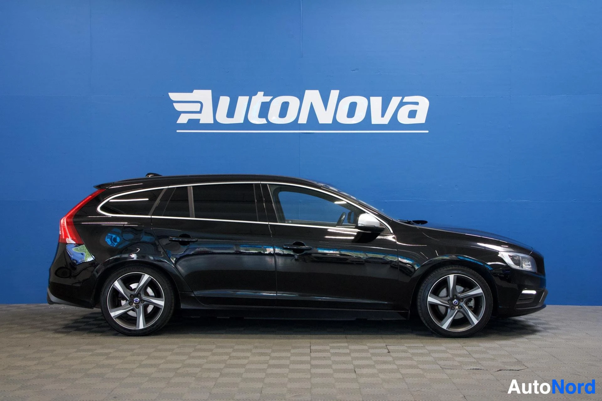 Etuviisto, nokka vasemmalle: Musta Volvo V60 etupuoli vasemmalle kääntyneenä, näkyvissä koko etupuoli ja vasen sivu, valkoiset vanteet, sininen seinä taustalla